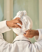 Person in a white robe adjusting a towel on their head against a neutral background